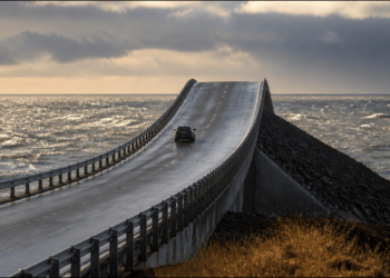 Carro trafega por uma ponte asfaltada sobre o mar, com pista elevada e curvada que parece terminar na água sob céu nublado.