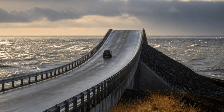 Carro trafega por uma ponte asfaltada sobre o mar, com pista elevada e curvada que parece terminar na água sob céu nublado.