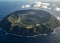 Vista aérea de Aogashima, uma ilha vulcânica cercada pelo oceano, com encostas íngremes, caldeira central coberta por vegetação e pequenas construções.
