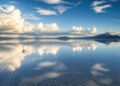 Vista ampla do Salar de Uyuni coberto por uma lâmina de água, refletindo nuvens e montanhas, com um veículo deixando marcas sobre a superfície.