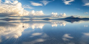 Vista ampla do Salar de Uyuni coberto por uma lâmina de água, refletindo nuvens e montanhas, com um veículo deixando marcas sobre a superfície.