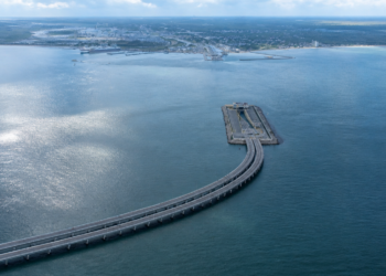 Vista aérea de uma extensa via elevada sobre o mar, conectada a uma plataforma retangular, com cidade costeira ao fundo.