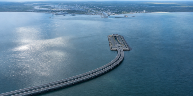 Vista aérea de uma extensa via elevada sobre o mar, conectada a uma plataforma retangular, com cidade costeira ao fundo.