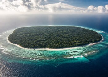 Vista aérea de uma ilha tropical coberta por floresta densa, cercada por praia clara, recifes e águas azul-turquesa.