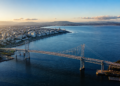 Vista aérea de uma grande ponte suspensa atravessando águas azuis, com área urbana à esquerda, montanhas ao fundo e céu claro ao entardecer.