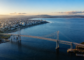 Vista aérea de uma grande ponte suspensa atravessando águas azuis, com área urbana à esquerda, montanhas ao fundo e céu claro ao entardecer.