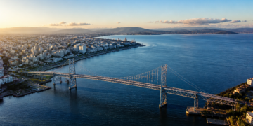 Vista aérea de uma grande ponte suspensa atravessando águas azuis, com área urbana à esquerda, montanhas ao fundo e céu claro ao entardecer.