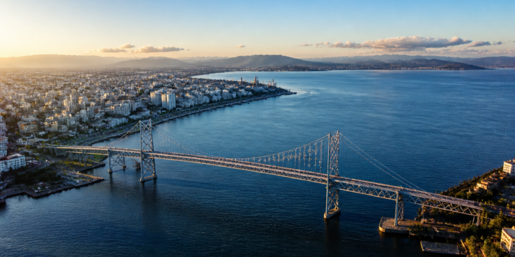 Vista aérea de uma grande ponte suspensa atravessando águas azuis, com área urbana à esquerda, montanhas ao fundo e céu claro ao entardecer.