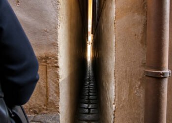Passagem extremamente estreita entre duas paredes, com piso de pedras, um cano lateral e parte de uma pessoa à esquerda.