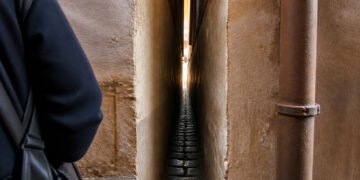 Passagem extremamente estreita entre duas paredes, com piso de pedras, um cano lateral e parte de uma pessoa à esquerda.