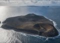 Vista aérea de uma ilha vulcânica escura cercada pelo oceano, com duas crateras bem definidas, litoral rochoso e pequenas ilhas ao fundo.