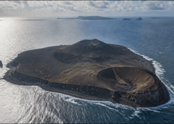 Vista aérea de uma ilha vulcânica escura cercada pelo oceano, com duas crateras bem definidas, litoral rochoso e pequenas ilhas ao fundo.