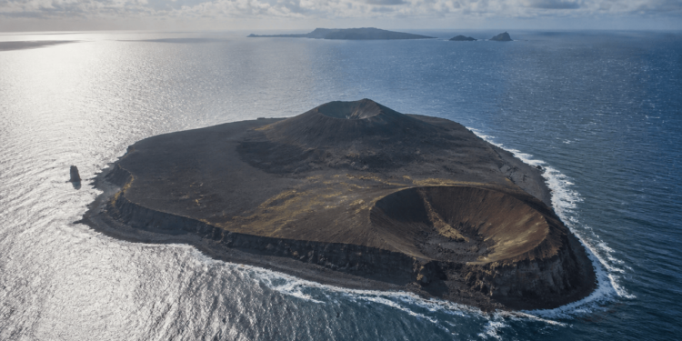 Vista aérea de uma ilha vulcânica escura cercada pelo oceano, com duas crateras bem definidas, litoral rochoso e pequenas ilhas ao fundo.