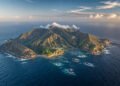 Vista aérea da Ilha da Trindade, com relevo montanhoso, costa rochosa, pequena área construída junto a uma enseada e oceano ao redor.