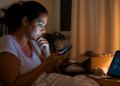Mulher sentada na cama à noite observa o celular com expressão preocupada, ao lado de uma mesa com abajur, caixa aberta e tablet com ícones na tela.