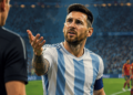 Jogador de futebol com uniforme listrado em azul e branco gesticula diante de um árbitro durante uma partida, com expressão de tensão e questionamento.