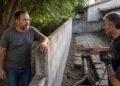 Homem encostado em um muro conversa com dois trabalhadores enquanto um encanamento exposto e raízes aparecem em uma vala ao lado, em área externa residencial.