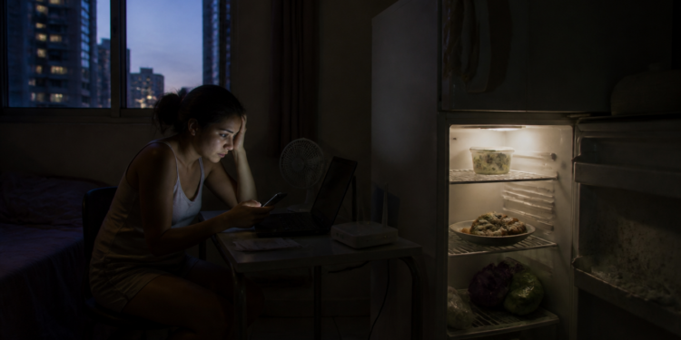 Jovem sentada diante de um notebook em uma kitnet escura, olhando o celular ao lado de uma geladeira aberta com alimentos armazenados.