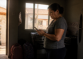 Mulher consulta o celular em uma cozinha escura enquanto um técnico uniformizado verifica o medidor de energia do lado de fora da residência.
