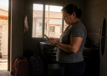 Mulher consulta o celular em uma cozinha escura enquanto um técnico uniformizado verifica o medidor de energia do lado de fora da residência.