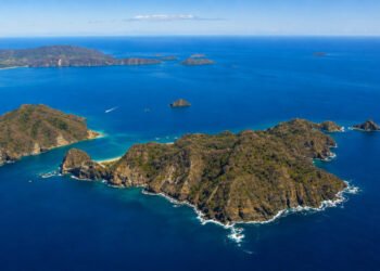 Vista aérea de ilhas montanhosas cercadas por mar azul, com enseadas de águas claras, pequenas praias e uma embarcação navegando entre as formações costeiras.