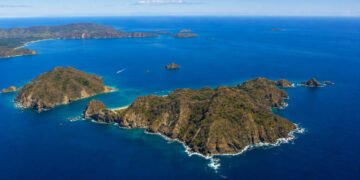 Vista aérea de ilhas montanhosas cercadas por mar azul, com enseadas de águas claras, pequenas praias e uma embarcação navegando entre as formações costeiras.