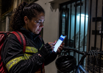 Entregadora com mochila vermelha e jaqueta com faixas refletivas consulta o celular à noite, ao lado de uma bicicleta e de um portão, em cena que remete a golpe por SMS sobre restituição.