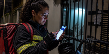 Entregadora com mochila vermelha e jaqueta com faixas refletivas consulta o celular à noite, ao lado de uma bicicleta e de um portão, em cena que remete a golpe por SMS sobre restituição.