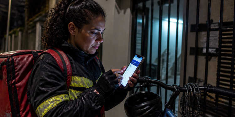 Entregadora com mochila vermelha e jaqueta com faixas refletivas consulta o celular à noite, ao lado de uma bicicleta e de um portão, em cena que remete a golpe por SMS sobre restituição.