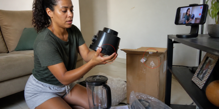 Mulher sentada no chão da sala segura a base de um liquidificador recém-retirado da caixa, enquanto grava a abertura com o celular e observa uma rachadura visível na peça.