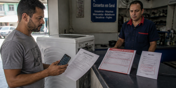 Homem em assistência técnica segura um celular e uma ordem de serviço ao lado de uma máquina de lavar, enquanto um atendente observa atrás do balcão com orçamento e recibo expostos.