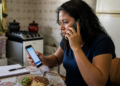 Mulher sentada à mesa da cozinha fala ao telefone enquanto consulta outro celular, com caderno de anotações e prato de comida à sua frente.