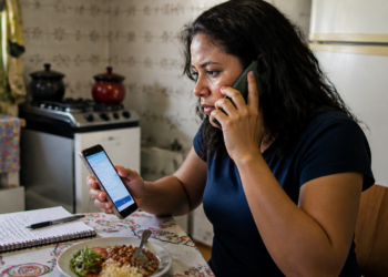 Mulher sentada à mesa da cozinha fala ao telefone enquanto consulta outro celular, com caderno de anotações e prato de comida à sua frente.