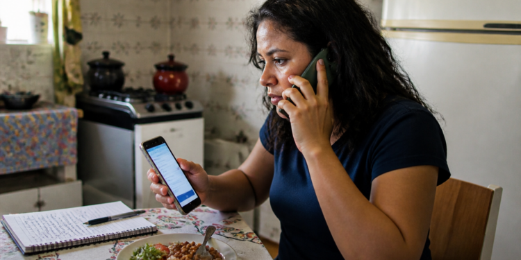 Mulher sentada à mesa da cozinha fala ao telefone enquanto consulta outro celular, com caderno de anotações e prato de comida à sua frente.