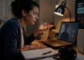 Mulher sentada à mesa em casa observa o computador durante uma videochamada com imagem travada, ao lado de caderno, fones, luminária e equipamento de internet.