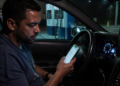 Homem sentado dentro de um carro à noite olha para o celular com expressão preocupada, com o painel iluminado e outro telefone preso ao suporte no painel.