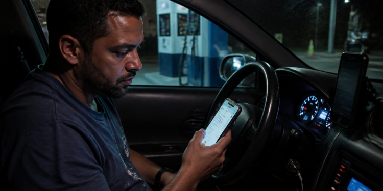 Homem sentado dentro de um carro à noite olha para o celular com expressão preocupada, com o painel iluminado e outro telefone preso ao suporte no painel.