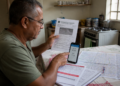 Homem sentado à mesa da cozinha segura uma notificação de trânsito e aponta para a tela do celular, ao lado de outros documentos, um mapa e um caderno.