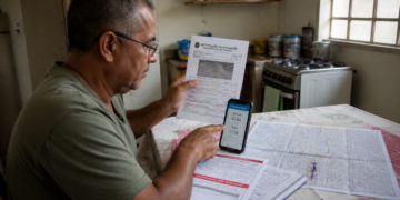 Homem sentado à mesa da cozinha segura uma notificação de trânsito e aponta para a tela do celular, ao lado de outros documentos, um mapa e um caderno.