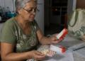 Mulher idosa sentada à mesa da cozinha observa uma caixa de remédio enquanto segura uma cartela com comprimidos de cores diferentes, com papéis e outra embalagem de medicamento sobre a mesa.