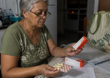 Mulher idosa sentada à mesa da cozinha observa uma caixa de remédio enquanto segura uma cartela com comprimidos de cores diferentes, com papéis e outra embalagem de medicamento sobre a mesa.