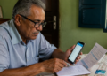 Homem idoso sentado à mesa compara informações exibidas no celular com um extrato impresso, enquanto faz anotações em um caderno ao lado de um cartão bancário.