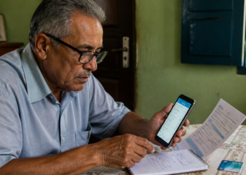 Homem idoso sentado à mesa compara informações exibidas no celular com um extrato impresso, enquanto faz anotações em um caderno ao lado de um cartão bancário.