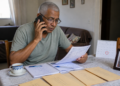 Homem idoso sentado à mesa fala ao telefone enquanto lê um documento, com caderno, envelopes, calendário e xícara ao redor, em cena de preocupação com pagamento e extrato.