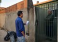 Homem com papéis conversa com um morador de braços cruzados atrás de um portão, ao lado de um muro parcialmente sem reboco e materiais de obra na calçada.