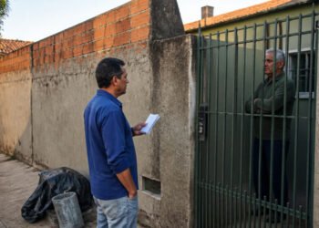 Homem com papéis conversa com um morador de braços cruzados atrás de um portão, ao lado de um muro parcialmente sem reboco e materiais de obra na calçada.