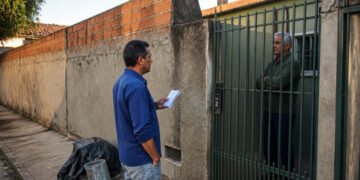 Homem com papéis conversa com um morador de braços cruzados atrás de um portão, ao lado de um muro parcialmente sem reboco e materiais de obra na calçada.