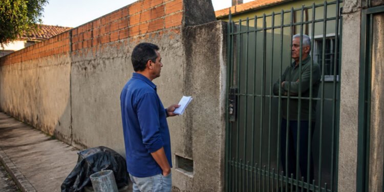 Homem com papéis conversa com um morador de braços cruzados atrás de um portão, ao lado de um muro parcialmente sem reboco e materiais de obra na calçada.