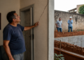 Homem aponta para uma rachadura na parede de casa enquanto, do lado de fora, dois homens observam uma escavação de obra ao lado do imóvel.