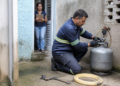 Técnico com uniforme azul e luvas verifica a conexão de um botijão de gás no quintal, enquanto uma moradora observa da porta segurando uma prancheta.